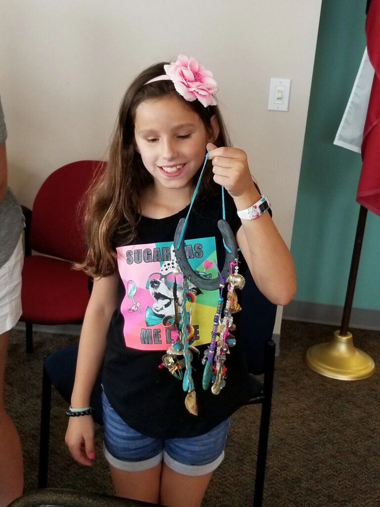 An artist holds up a finished horseshoe necklace hung with colourful beaded strands, small charms and a shell.