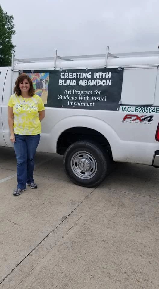 A volunteer stands beside a pickup truck reading Creating With Blind Abandon, Art Program for Students With Visual Impairment, printed on the truck's rear panel.