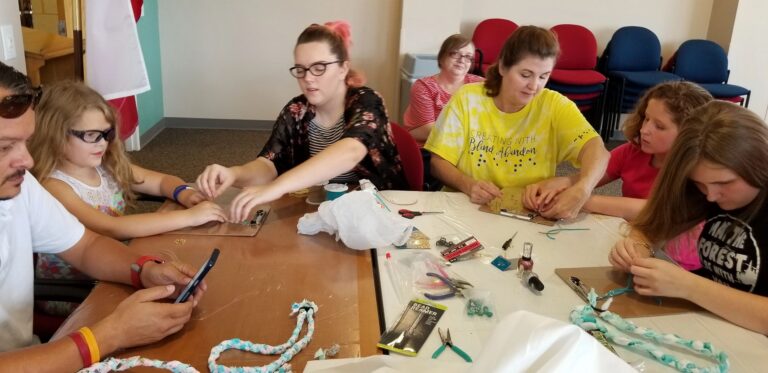 Several artists and volunteers gather around a table braiding fabric and beading, with pliers, nail polish and craft supplies spread across the surface.