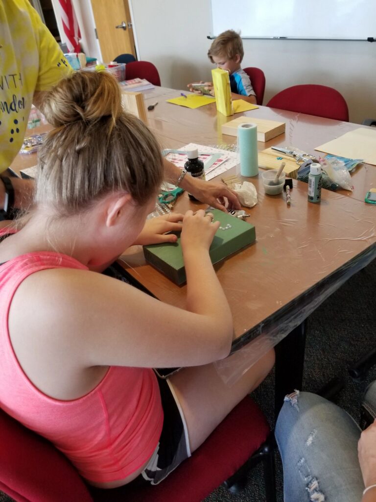 An artist decorates a green box with rhinestones at a table set with paint, ribbon and craft supplies.