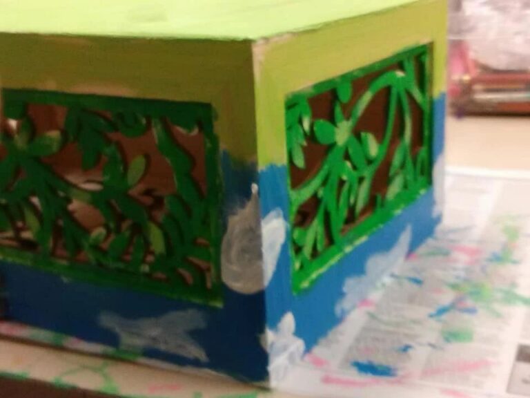 A wooden box with a carved leaf-and-vine cutout pattern, painted in layers of blue and green, resting on a table covered in newspaper.