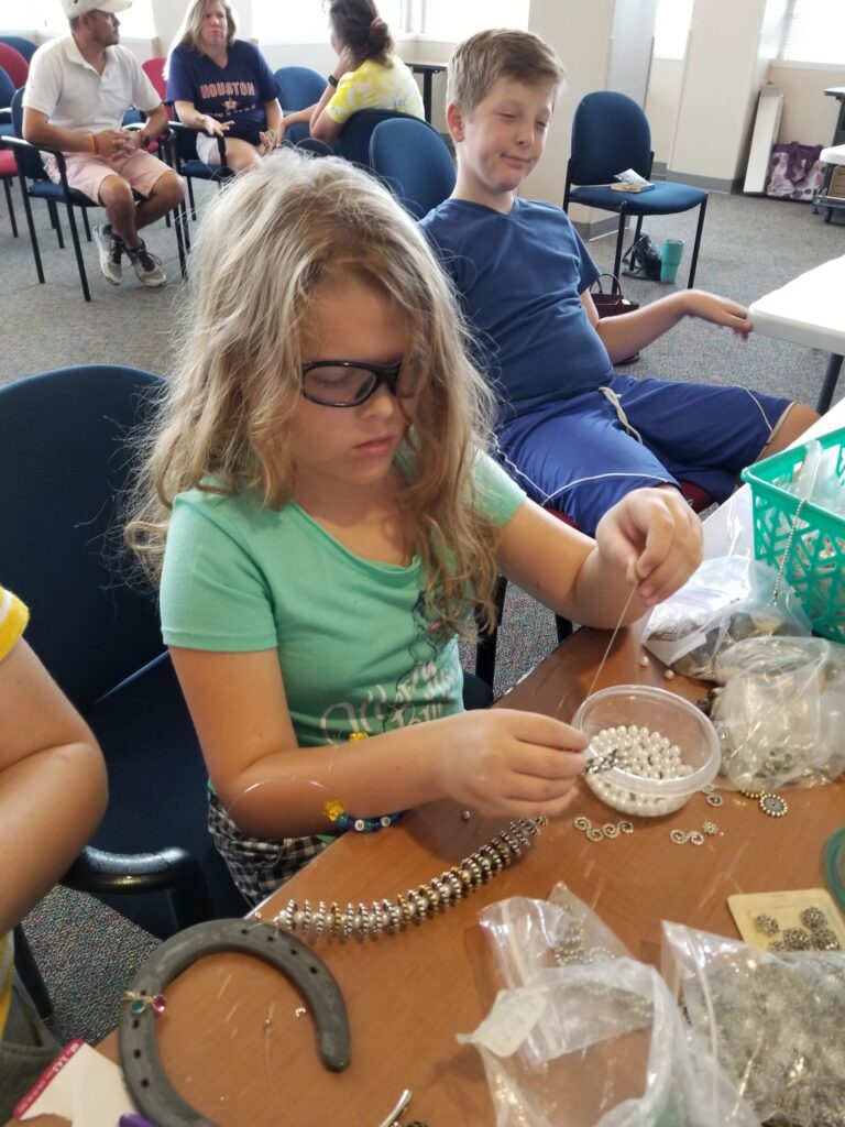 An artist threads pearl beads onto wire at a table with a horseshoe and a beaded bracelet nearby.