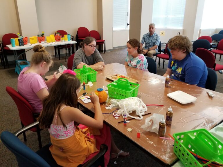 Artists sit around a table decorating a small pumpkin with paint and glue, yarn and craft supplies spread across the surface.