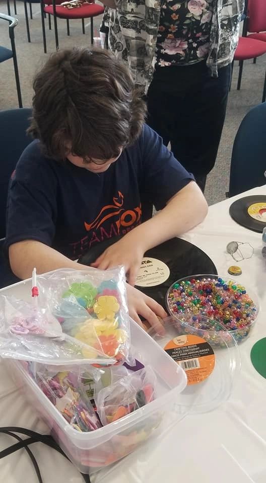 An artist reaches into a bin of beads and foam flower shapes at a table set with a decorated record and a bowl of beads.