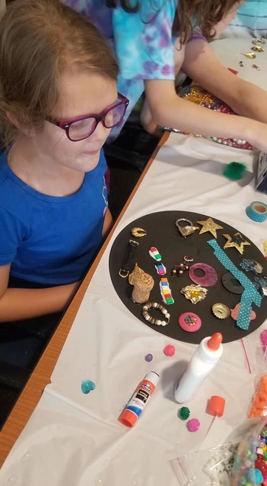 An artist looks over a black display board covered in pins, charms, a cork and small trinkets, glue supplies nearby on the table.