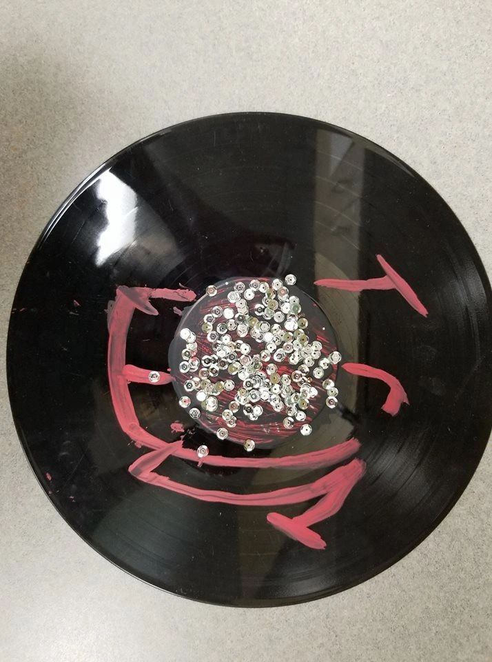 A black record with a red arrow pattern and a cluster of silver sequins pressed into wet paint at the centre.