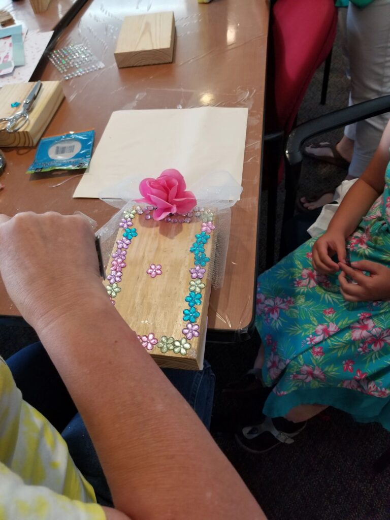 A small wooden box decorated with a border of flower gems, resting on a table beside a pink fabric rose.