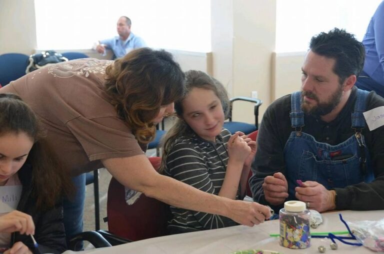 A volunteer helps an artist thread beads at a table set with a jar of beads, colourful pipe cleaners and other artists working nearby.