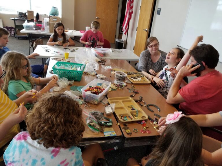 A group of artists and volunteers gather around a table covered in horseshoes, beads and jewellery supplies.