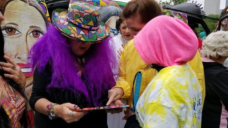 A person in a purple wig and hand-painted hat looks closely at a beaded braille tag while others in tie-dye and yellow jackets gather around a painted parade car.