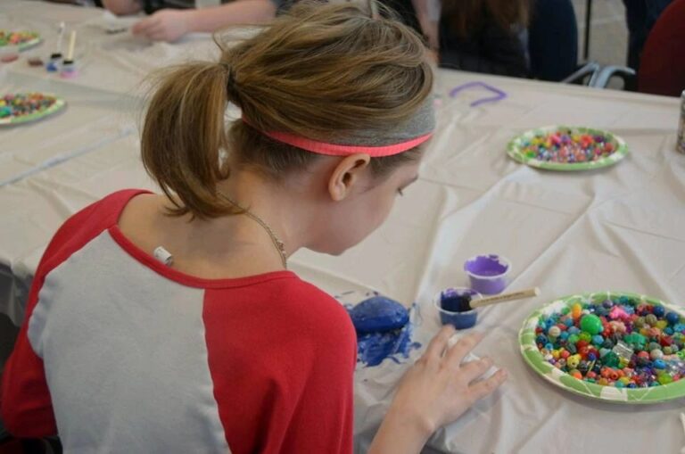 An artist paints a stone bright blue with a brush at a table set with cups of beads and paint.