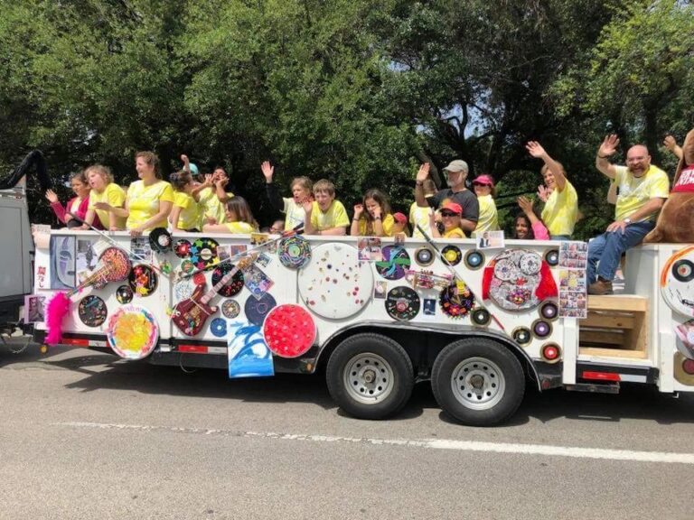 The float parked on the parade route, riders waving from the platform and the truck's raised perch, downtown high-rises visible in the background.