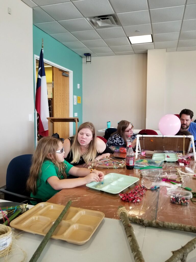 A group of artists and volunteers sit at a table weaving yarn onto stick-frame dream catchers, beads and craft supplies spread across the surface.
