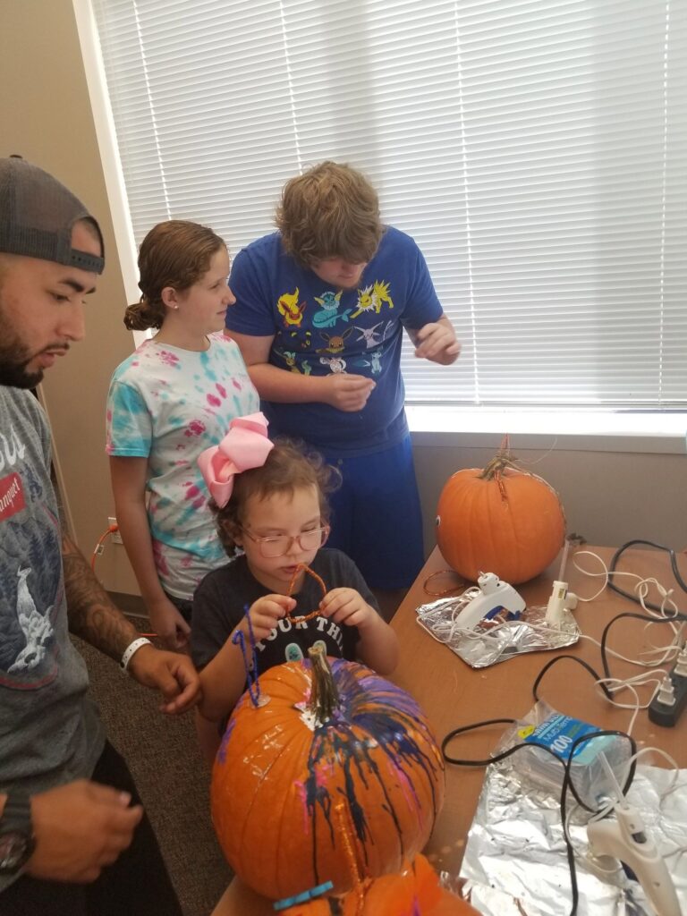 Artists decorate pumpkins with hot glue guns at a table, splattering one pumpkin with purple and orange paint.