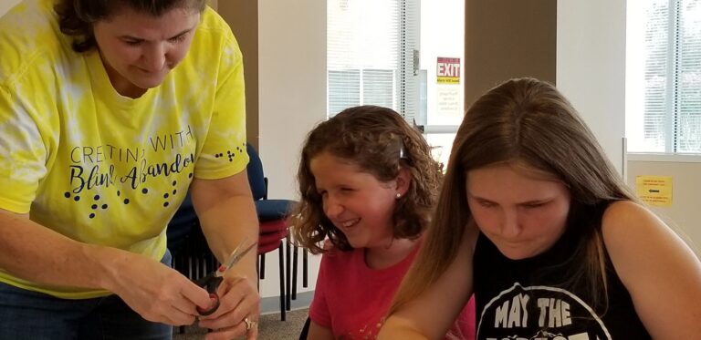 A volunteer trims a beaded cord with scissors for an artist, another artist smiling beside them at the table.