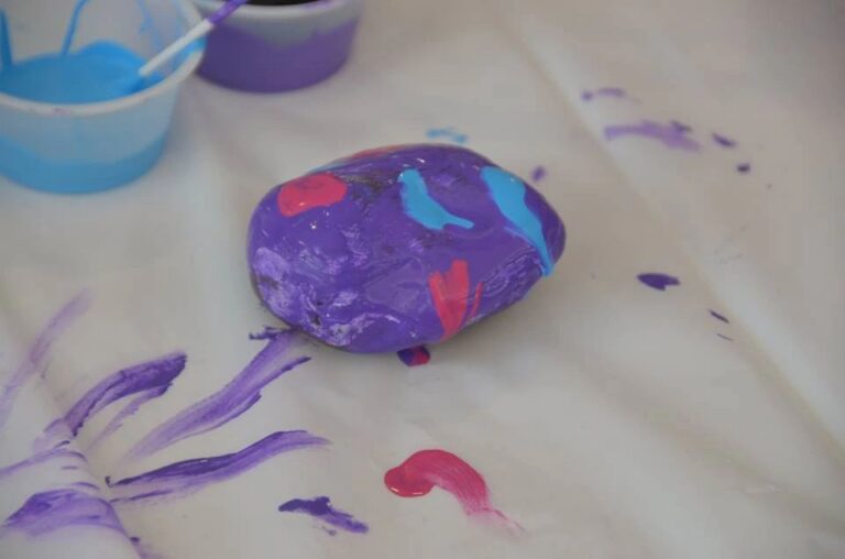 A stone painted white with a bright teal flower pattern dabbed on using a sponge, resting beside a cup of teal paint.