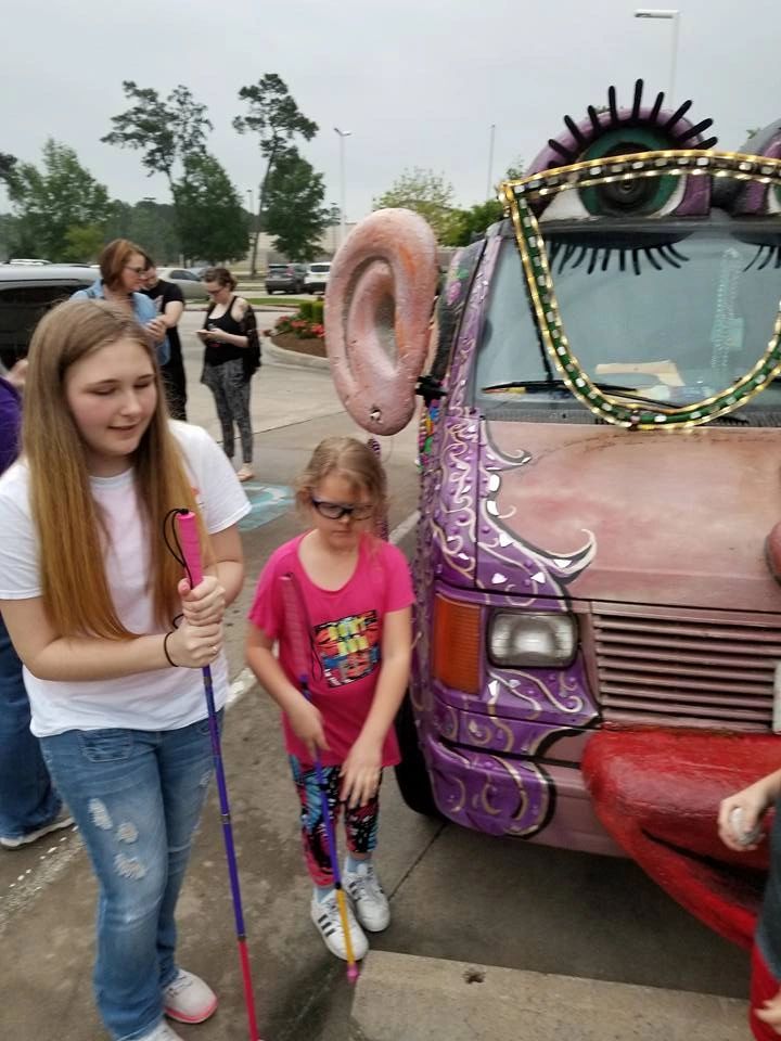 Two young artists holding white canes stand beside the front of the Women Rock Art Car, its large sculpted ear and painted lashes visible above them.