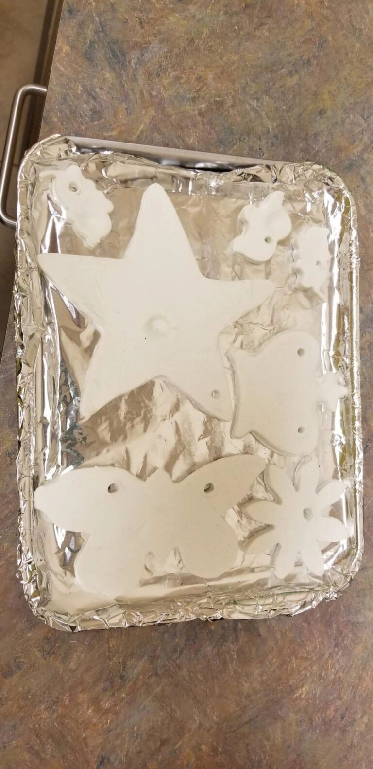 A foil tray holding several dry-brushed white dough ornaments shaped as a star, flowers, a bunny and a bird, ready for painting.