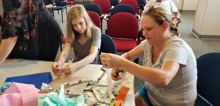 An artist and a volunteer braid pastel fabric strips together, with more braided ropes coiled on the table beside them.