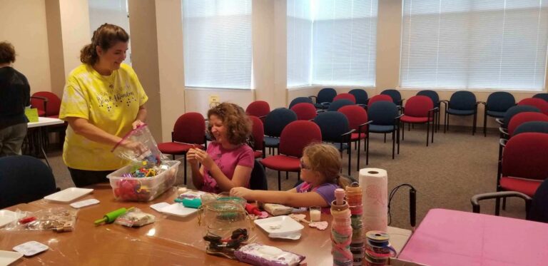 A volunteer shows a bin of small charms to two artists at a table set with rolling pins and dough shapes.