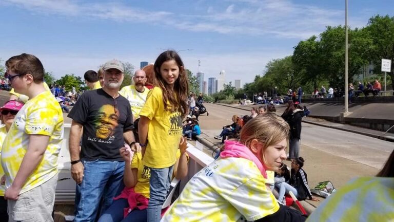 A group stands together on the float overlooking downtown, one wearing a t-shirt printed with a portrait, everyone smiling for the photo.