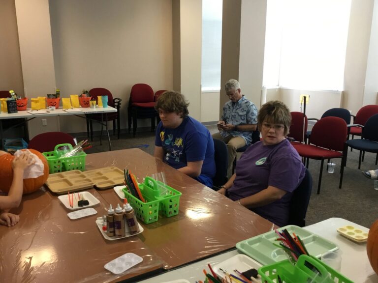 A group of artists and volunteers sit at a table with pumpkins and craft supplies, small pumpkin-themed treat buckets on a side table.