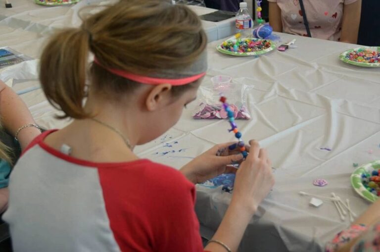 An artist paints a small blue bead at a table, with plates of colourful beads and craft supplies spread around them.