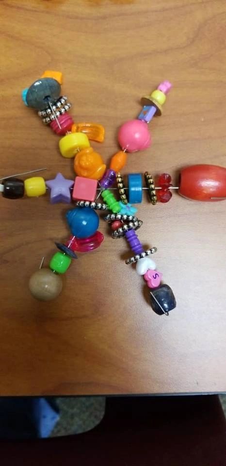 A beaded figure with arms and legs of colourful beads radiating from a central bead, a stone at one end and small charms scattered throughout.