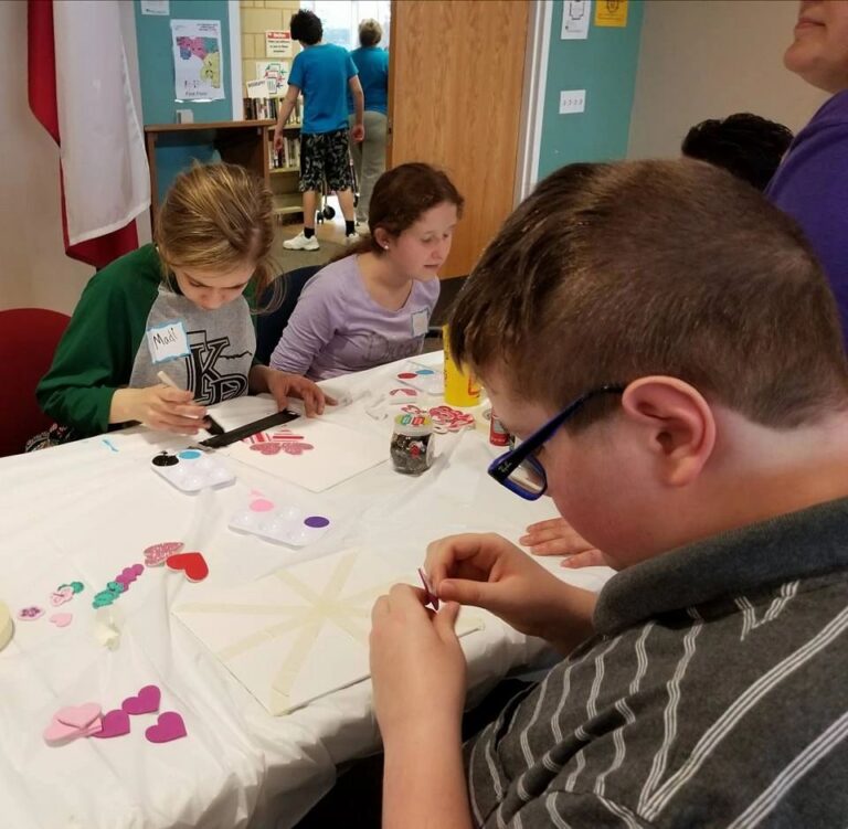 Two artists work on Valentine-themed canvases at a table covered with paint palettes, heart stickers and craft supplies, a volunteer nearby.