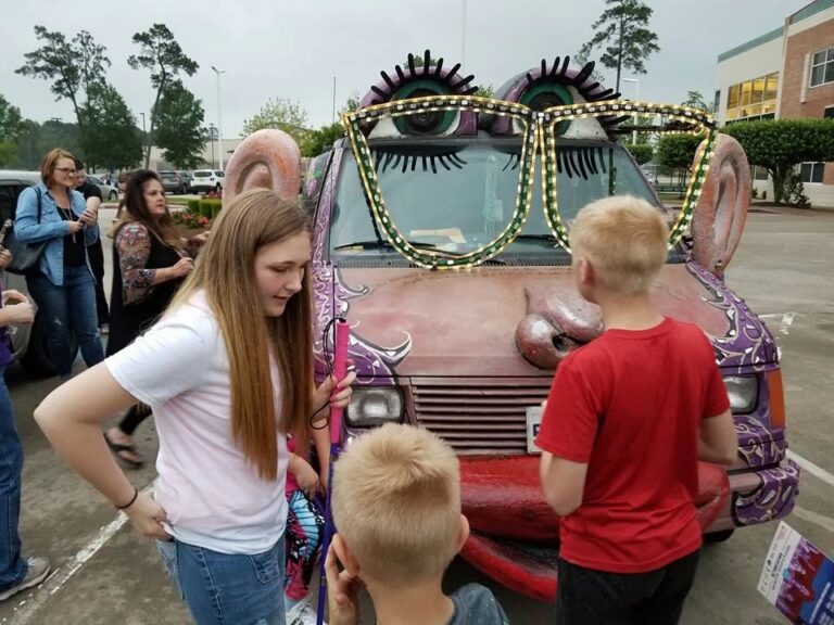 Three young artists gather around the front of the Women Rock Art Car, its lash-lined eyes, sculpted ear and painted nose visible above them.