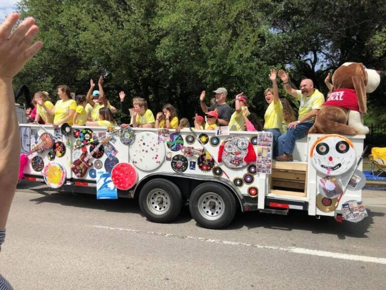 The float travels down the parade route past trees and buildings, riders waving from both the float and the truck's raised platform.