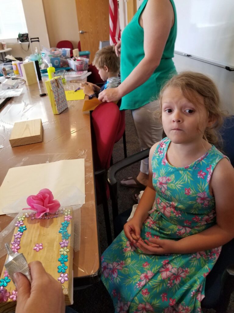 An artist sits at a table where another person decorates a wooden box with a pink fabric rose and flower gems.