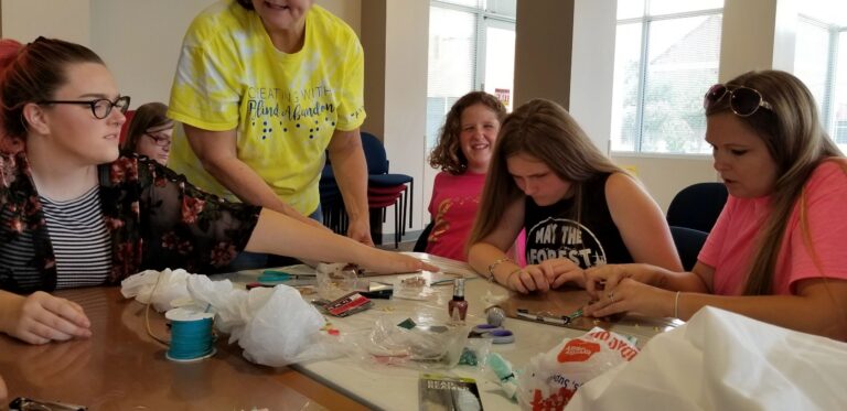 A volunteer helps a group of artists bead at a table covered in thread spools, plastic bags and craft supplies.