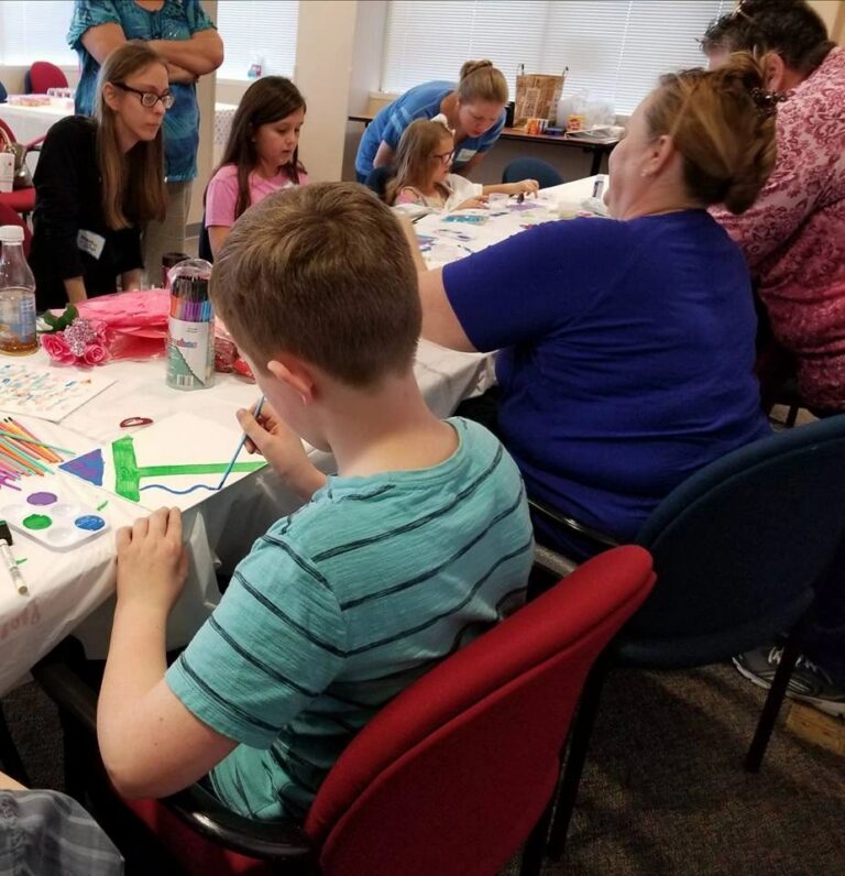 A wide view of the studio, with artists and volunteers working on Valentine-themed canvases at a long table covered in paint palettes and craft supplies.