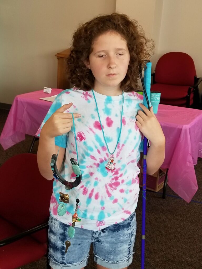 An artist stands holding a beaded necklace with a horseshoe pendant, a white cane resting against their shoulder.