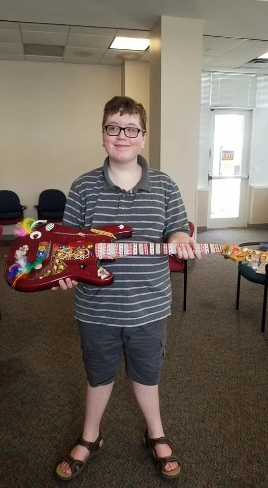 An artist holds up a red electric guitar decorated with feathers, gems and small charms along its body.