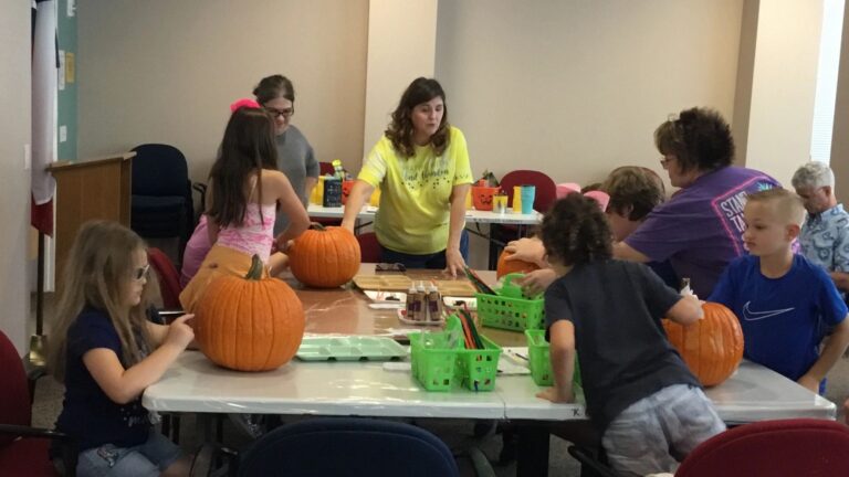 A group of artists and a volunteer work at a table covered in whole pumpkins and craft trays.