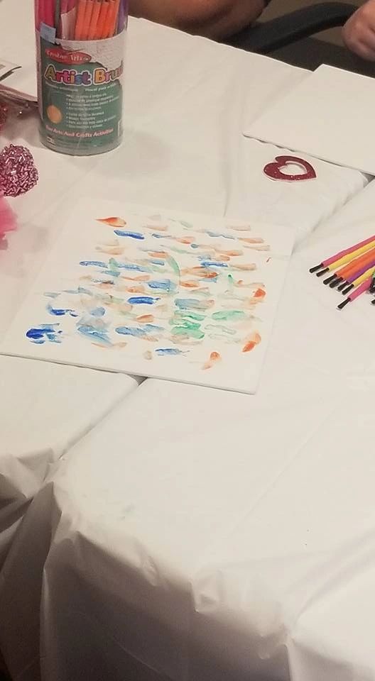 A canvas decorated with a large blue circle covered in shiny black gem buttons, bordered by a pink pipe cleaner, with a red glitter flower painted in the corner.