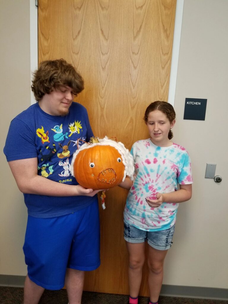 Two artists hold up a pumpkin wrapped in white yarn with googly eyes and gold-studded accents.