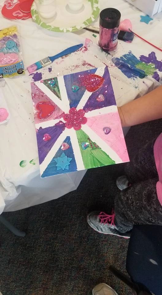 A finished canvas divided into coloured triangles in blue, pink, purple and green, decorated with glitter, gem hearts and a glittery flower at the centre, held up by an artist.
