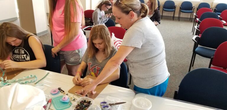 A volunteer helps a young artist bead on a clipboard while two more artists work beside them, braided cord and beads scattered on the table.