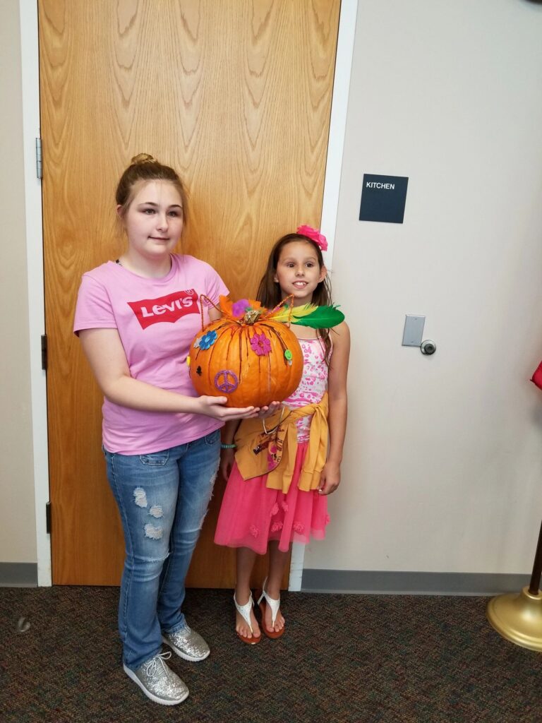 Two artists hold up a pumpkin decorated with a peace sign, painted flowers and a purple ribbon around its stem.
