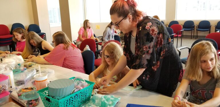 A volunteer helps two artists braid fabric at a table, with turquoise storage bins and bags of ribbon nearby.