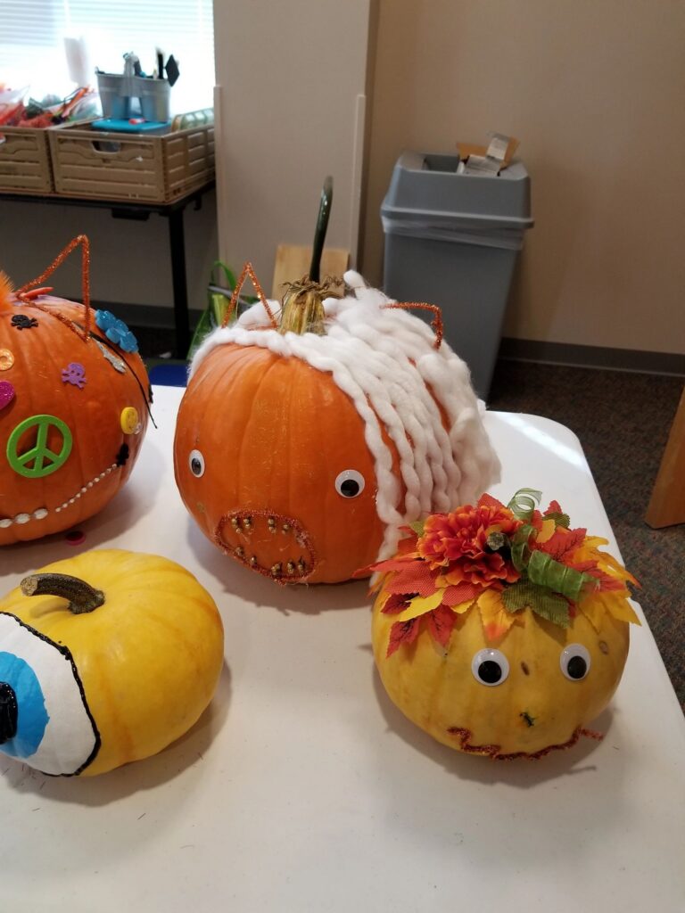 Four decorated pumpkins on a table: one with a peace sign and pastel gems, one wrapped in white yarn with googly eyes and gold studs, and two small yellow gourds painted as faces.