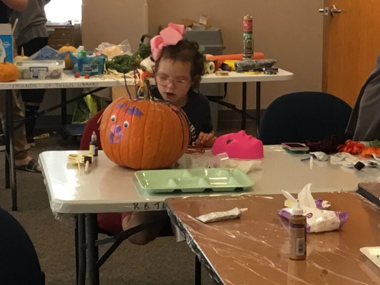 An artist sits at a table with a pumpkin painted as a face, glue and craft supplies nearby.