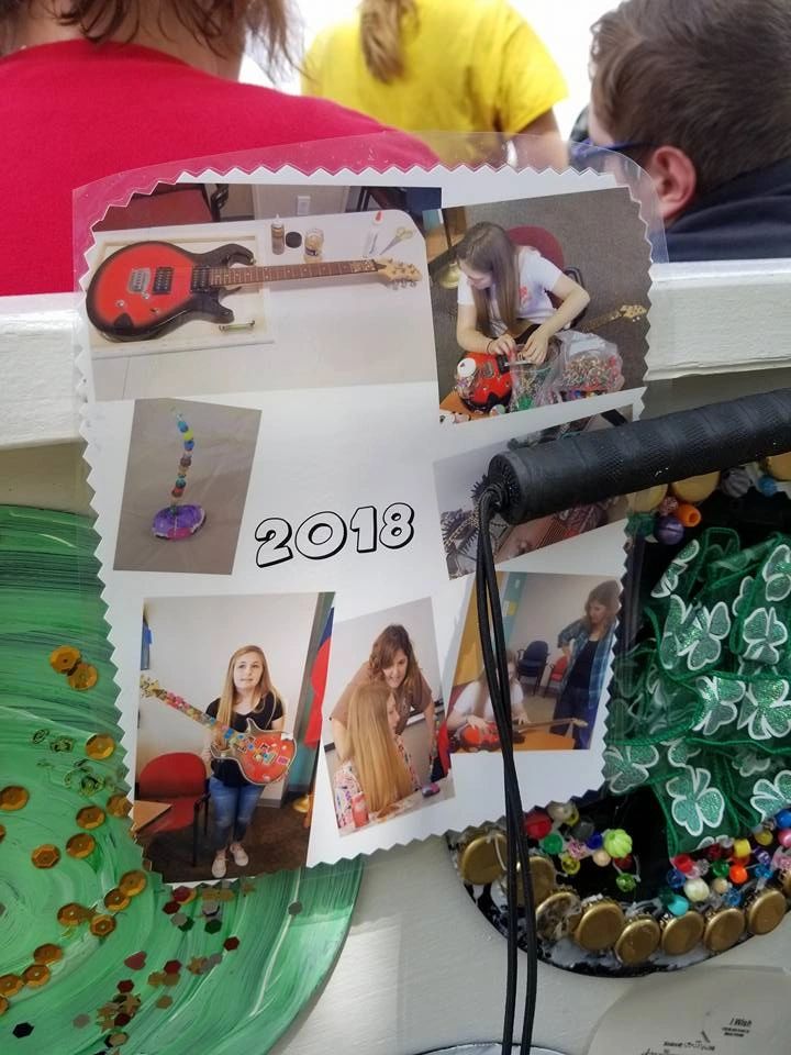 A laminated card labelled 2018, showing photographs of past projects: a guitar, a beaded stone, decorated records, and artists at work.