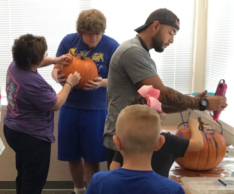 A volunteer helps two artists paint a pumpkin at a table, another artist working on a second pumpkin nearby.