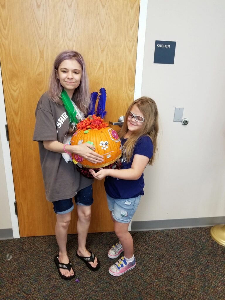 Two artists hold up a pumpkin decorated with painted flowers, feathers around the stem, and a face made from googly eyes and glitter pipe cleaners.