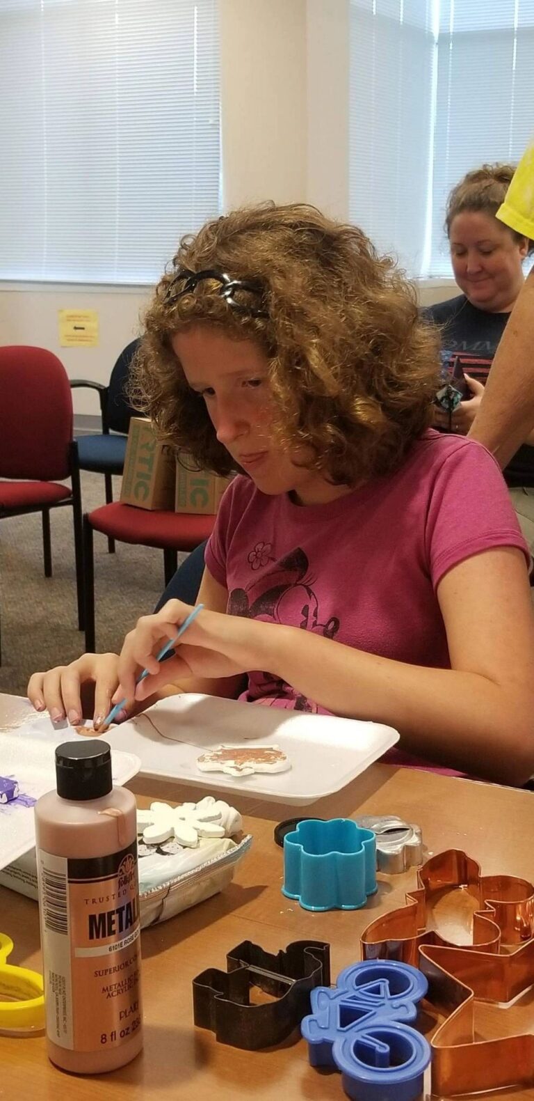An artist paints a dough ornament with metallic rose paint, cookie cutters shaped as flowers, stars and a bicycle spread nearby.