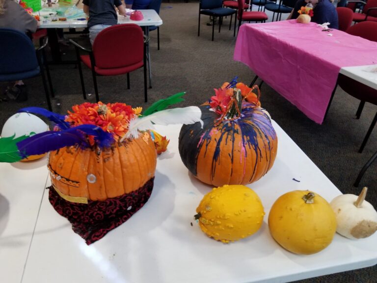 Two decorated pumpkins on a table: one with a feathered floral top and painted face, the other splattered with purple and blue paint, alongside small painted gourds.